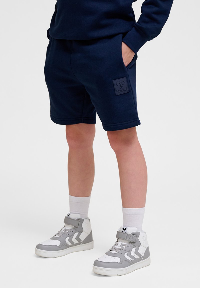 Navyblauwe katoenen shorts met zijzakken en gegraveerde logopatch. Draagt bij grijs-witte hoge sneakers en witte crew socks.