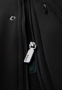 Tejido de chaqueta negra con un cierre metálico plateado y una pestaña en forma de hexágono grabada con "ombre". Primer plano del detalle de la costura.