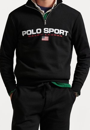 Uomo con felpa nera con zip a quarto e scritta "POLO SPORT RALPH LAUREN" con una bandiera americana, pantaloni neri e mani nelle tasche.