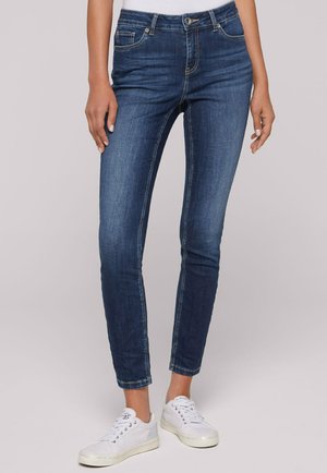 Dark blue, slim-fit jeans with a mid-rise waist, featuring five pockets and contrasting stitching. Paired with white sneakers.