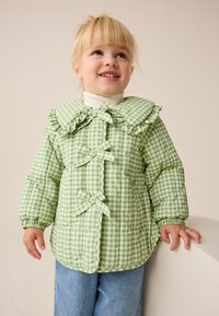 Grüne, kariert gesteppte Jacke mit voluminösem Schnitt, einem gerafften Kragen und Schleifendetails. Verfügt über lange Ärmel und einen abgerundeten Saum, getragen über einem weißen Rollkragenpullover.