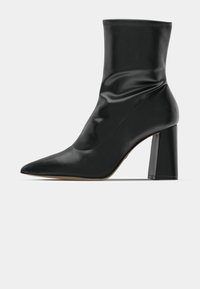 Black ankle boot made of shiny material, featuring a pointed toe and a chunky square heel. Smooth texture with gathered details near the ankle.