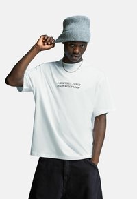 Jonge man met een grijze gebreide beanie, een wit t-shirt met de tekst "EEN MOOIE FOUT IN EEN PERFECTE LOOP," zwarte shorts, die de rand van zijn hoed aanraakt.