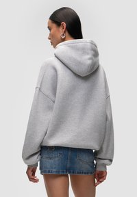 Hoodie grigio oversize con cappuccio, spalle abbassate e polsini a costine, abbinato a una mini gonna di jeans. Il tessuto appare morbido con una texture liscia.
