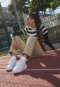 Sneakers bianchi, jeans beige a gamba larga, maglietta nera a maniche lunghe, camicia a righe a maniche corte; seduto contro una recinzione su una superficie rossa.