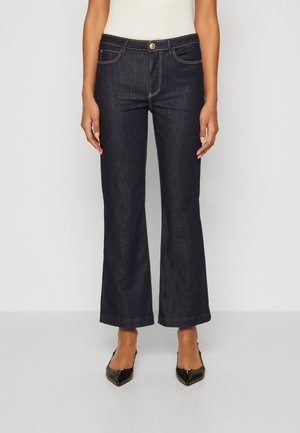 Van taille tot vloer zicht op een persoon die donkerblauwe bootcut jeans, een beige top en zwarte puntige hakken draagt tegen een effen achtergrond.