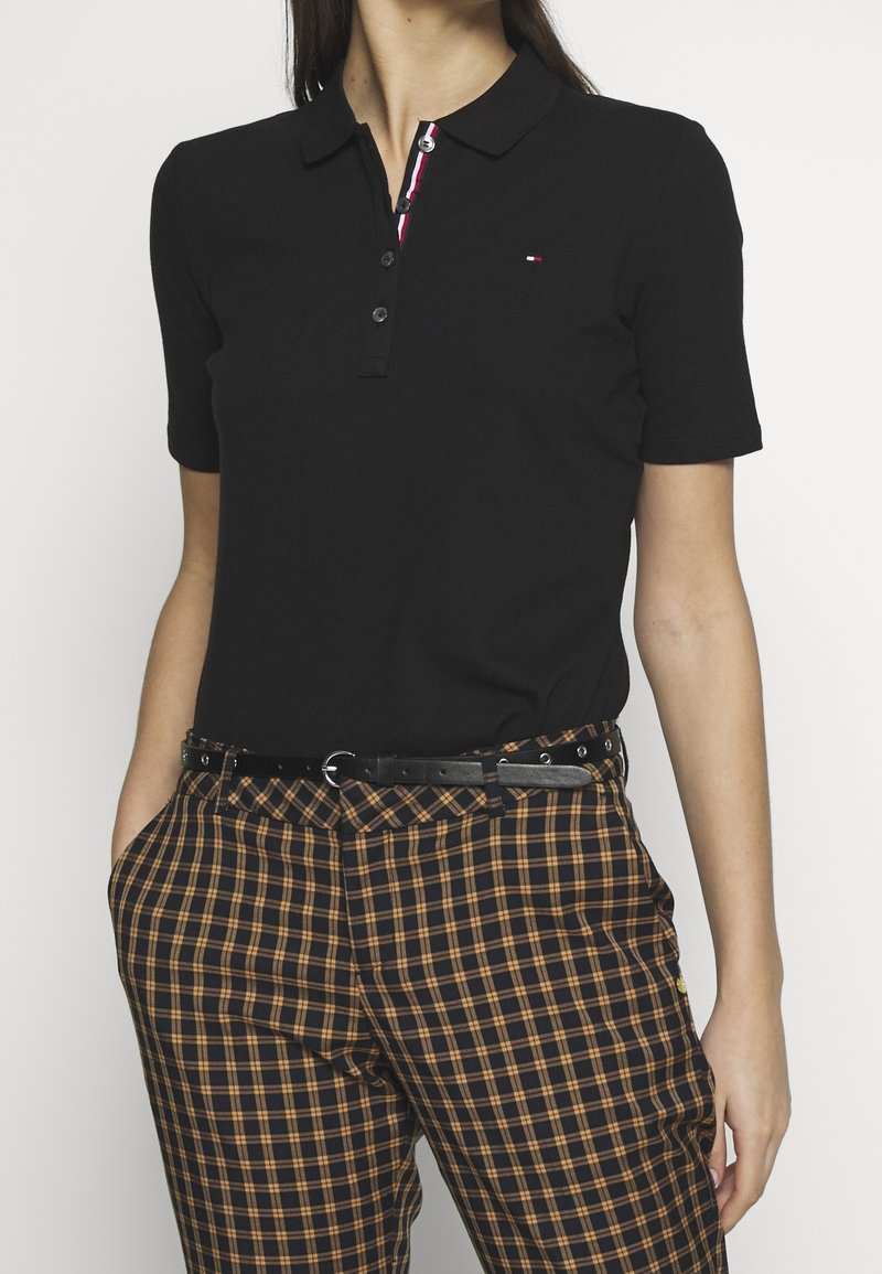 Femme portant un polo noir à manches courtes avec boutons et un pantalon à carreaux orange et noir avec une ceinture noire.
