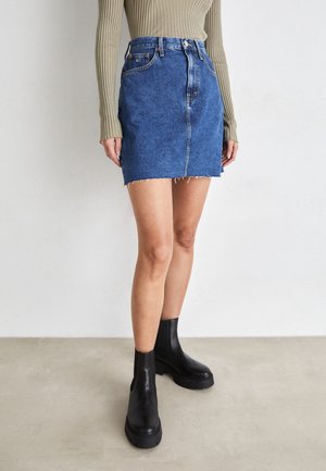 Personne portant une mini-jupe en jean bleu effilochée, un haut beige en maille côtelée à manches longues et des bottines noires épaisses, debout sur un sol gris.