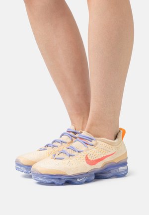 Des chaussures Nike Air VaporMax avec un dessus en maille beige clair, des accents orange et rouge, et une semelle de coussin transparent. Elles sont dotées de lacets bleus.
