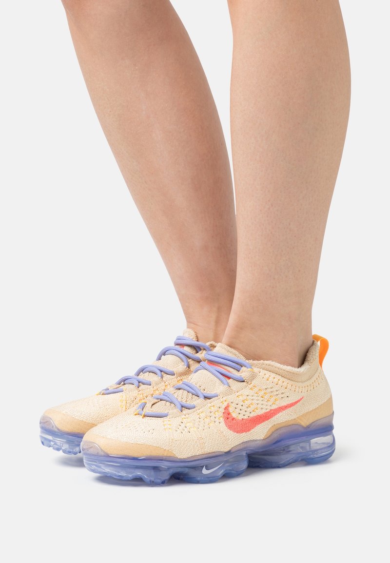 Nike Air VaporMax-skor med en ljusbeige stickad ovandel, orange och röda detaljer samt genomskinlig dämpningssula. Utrustad med blå snören.