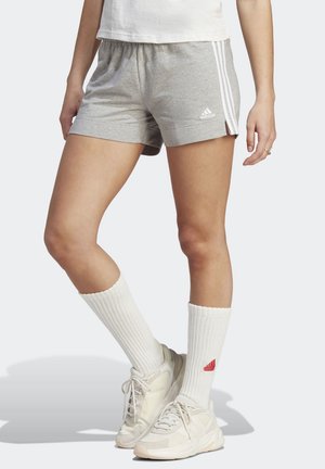 Person wearing grey Adidas shorts with white stripes, white crew socks with red logo, and off-white athletic shoes, standing on a light background.