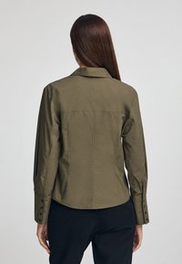 Olivgrön skjorta med knappar och långa ärmar, en spetsig krage och en figurnära design. Har synliga sömmar och knappdetaljer på ärmarna.