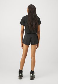 Svart, avkortet t-skjorte og shorts med en jevn tekstur. Båret med tursko og svarte sokker, sett fra baksiden.