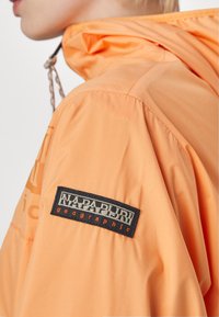 Pomarańczowa, lekka kurtka z kapturem, posiadająca fakturowany materiał oraz czarną metkę na rękawie z białym i pomarańczowym tekstem.