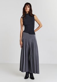 Donna che indossa un top a maniche corte nero a costine, pantaloni palazzo grigi plissettati a vita alta e stivaletti neri con tacco alto, in piedi su uno sfondo bianco.