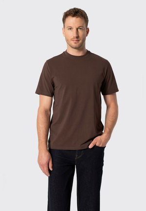 Hombre con cabello corto y rizado que lleva una camiseta marrón lisa de manga corta y vaqueros oscuros, con una mano en el bolsillo, de pie frente a un fondo claro.