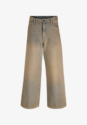 Tienerjongen staand, gekleed in een licht beige T-shirt met korte mouwen, losse vervaagde spijkerbroek en witte sneakers, kijkt schuin weg tegen een effen achtergrond.