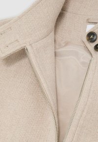 Beige Reißverschlussjacke mit einer strukturierten Außenseite und einem glatten Innenfutter, ausgestattet mit einem hohen Kragen und dunklen Knöpfen an der Vorderseite.