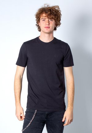 T-shirt nera a girocollo realizzata in tessuto morbido, con maniche corte e un piccolo logo sul petto, abbinata a jeans scuri e una catena.