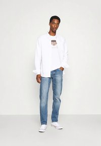 Cămașă albă cu mâneci lungi peste un tricou alb cu grafică cu un design de scut, asortată cu blugi deschis la culoare și adidași albi.