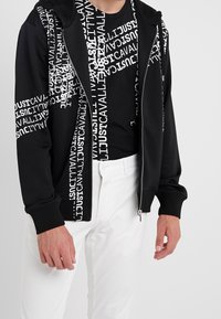 Czarna hoodie na zamek z białym napisem, w połączeniu z czarnym T-shirtem z podobnym napisem. Białe spodnie dopełniają strój.