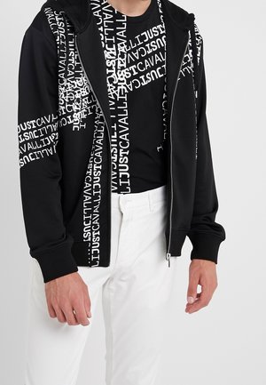 Schwarze Zip-Hoodie mit weißem Textdesign, kombiniert mit einem schwarzen T-Shirt mit ähnlichem Text. Weiße Hose rundet das Outfit ab.