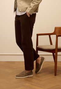 Pantalons en velours côtelé marron, chaussures beiges à enfiler avec semelles blanches, et une veste beige clair, se tenant à côté d'une chaise en bois avec un siège en tissu.