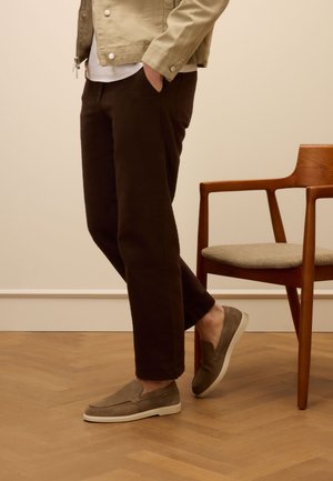 Braune Cordhosen, beige Slipper mit weißen Sohlen und eine hellbeige Jacke, stehen neben einem Holzstuhl mit Stoffsitz.