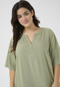 Blusa verde claro de manga corta con escote en V y pequeño detalle de muesca. Elaborada con una tela suave y lisa con un corte relajado.