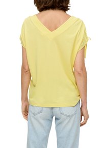 Gelbes V-Ausschnitt-T-Shirt mit kurzen Ärmeln und umgeschlagenen Ärmeln, lässiger Schnitt und glatter Textur. Getragen mit hellblauen Jeans.