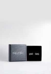 Silver infinityörhängen visade i en svart sammetssmyckeslåda med en texturerad svart yta och vit logotyp på framsidan.