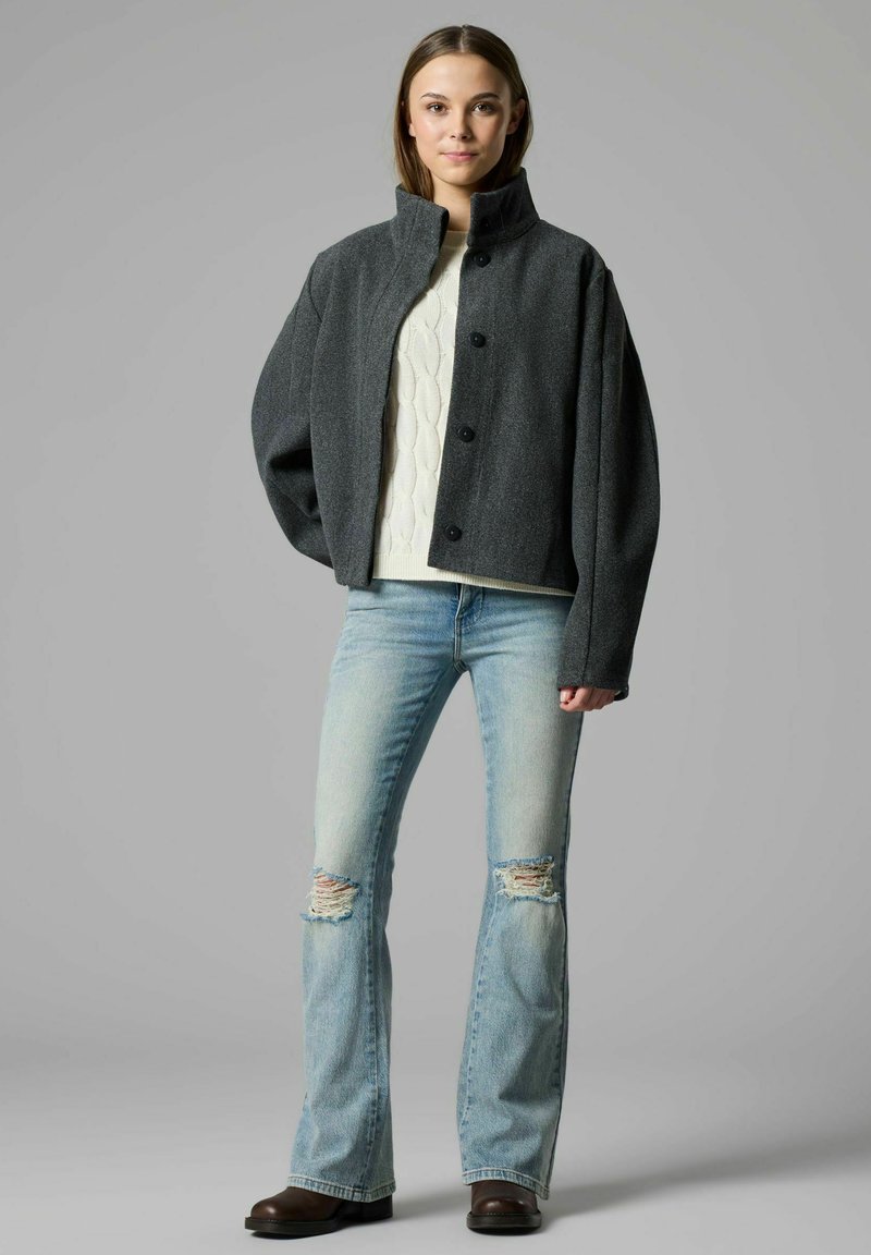 Jeune femme portant une veste boutonnée gris foncé, un pull en maille torsadée blanc, un jean bleu clair déchiré et des chaussures marron, debout devant un fond gris.