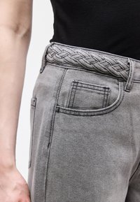 Κοντινό πλάνο ενός ατόμου που φοράει ανοιχτό γκρι τζιν παντελόνι με πλεκτή ζώνη και μαύρο μπλουζάκι που έχει μέσα στην μέση, εμφανίζοντας τη μπροστινή τσέπη και τα θηλύκια της ζώνης.