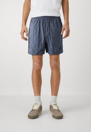 Homme portant un short Adidas bleu marine, des chaussettes blanches, des chaussures décontractées beiges, et un t-shirt blanc, debout devant un fond uni.