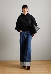 Black hoodie with drawstrings, paired with wide-legged denim jeans featuring a light blue hem. Model holds a black leather clutch.