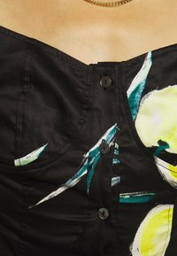 Indumento nero con bottoni e stampa floreale verde e gialla sulla parte alta del petto di una persona, che indossa una collana d'oro.