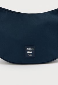 Torba tamnoplave boje s zakrivljenim oblikom, izrađena od izdržljive tkanine, s pravokutnom logotipskom oznakom na kojoj piše "LACOSTE PARIS." Zatvaranje na patentni zatvarač.
