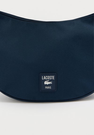 Sac bleu marine de forme courbée, fabriqué en tissu durable, doté d'un patch logo rectangulaire portant l'inscription "LACOSTE PARIS". Fermeture éclair.