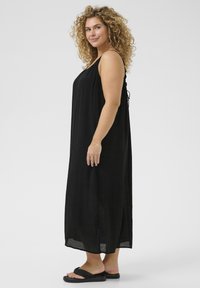 Vestido maxi negro con tirantes finos, material ligero, silueta fluida y aberturas laterales. Combinado con chanclas negras.