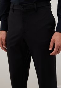 Uomo che indossa pantaloni neri e una camicia scura a maniche lunghe, in piedi con le mani rilassate ai fianchi davanti a uno sfondo semplice.