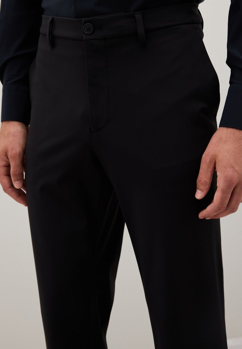 Uomo che indossa pantaloni neri e una camicia scura a maniche lunghe, in piedi con le mani rilassate ai fianchi davanti a uno sfondo semplice.