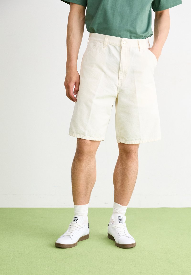 Shorts en coton beige clair avec une coupe droite, dotés de poches latérales et d'une fermeture à bouton, associés à des baskets blanches et une chemise verte.