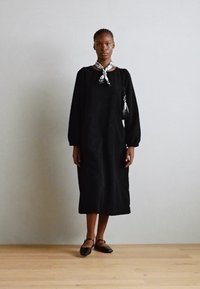 Čierne šaty s dlhým rukávom, zaobleným výstrihom a textúrovanou látkou. Na krku je viazaný svetlý šál. Outfit dopĺňajú čierne baleríny a malá kabelka.