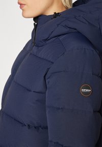 Tumši zils pūšļu mētelis ar kapuci, quilted dizainu un apaļu logo noņēmēju kreisajā piedurknē. Audums izskatās gluds ar matētu apdari.