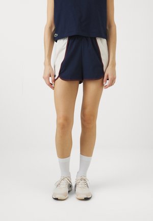 Persoon draagt marineblauwe en witte sportshorts met bordeaux afwerking, marineblauw shirt, witte sokken en beige hardloopschoenen, staand tegen een effen achtergrond.