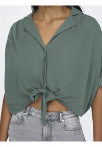 Groene, lichtgewicht, cropped blouse met een knoopsluiting aan de voorkant, korte mouwen en een strikdetail aan de voorkant. Gecombineerd met zilverkleurige spijkerbroek.
