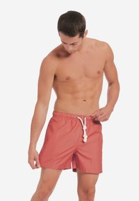 Punainen uima-asu, jossa on joustava vyötärö, valkoinen nauha ja lyhyet lahkeet. Suora pinta, minimalistinen muotoilu, ei näkyviä kuvioita tai koristeita.