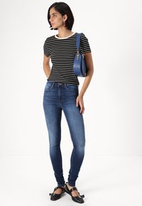 Črno-bela prugasta majica s krožnim izrezom, v kombinaciji s temno modrimi oprijetimi kavbojkami. Model nosi okroglo modro torbico in črne balerinke.