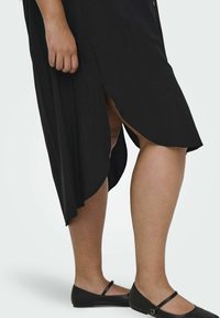 Robe noire avec une fente sur le côté, fabriquée dans un tissu doux. Une main saisit le tissu ; elle porte des chaussures plates noires avec un détail de bride.