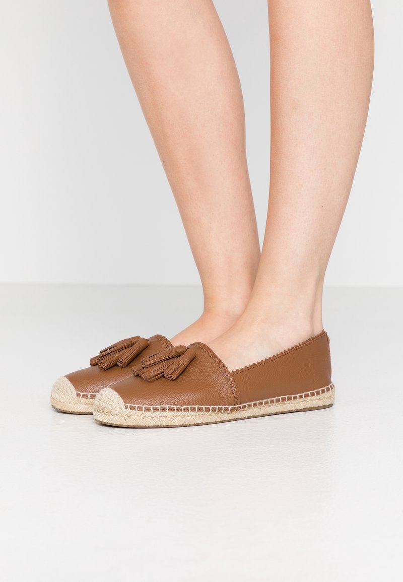 Espadrilles plates en cuir marron avec pompons portées pieds nus sur un fond clair uni.